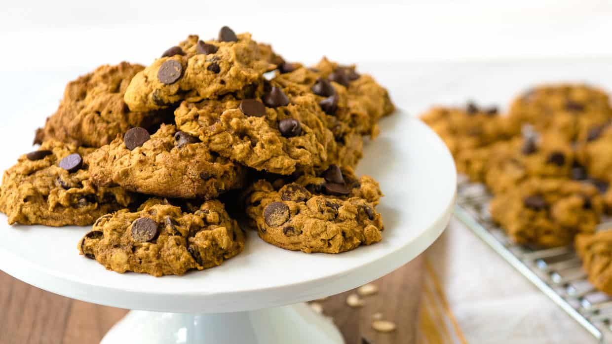 Pile of pumpkin oatmeal chocolate chip cookies with more in the background on a cooling rack.