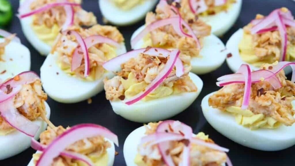 Deviled eggs topped with shredded meat and thinly sliced red onions are arranged on a black plate.