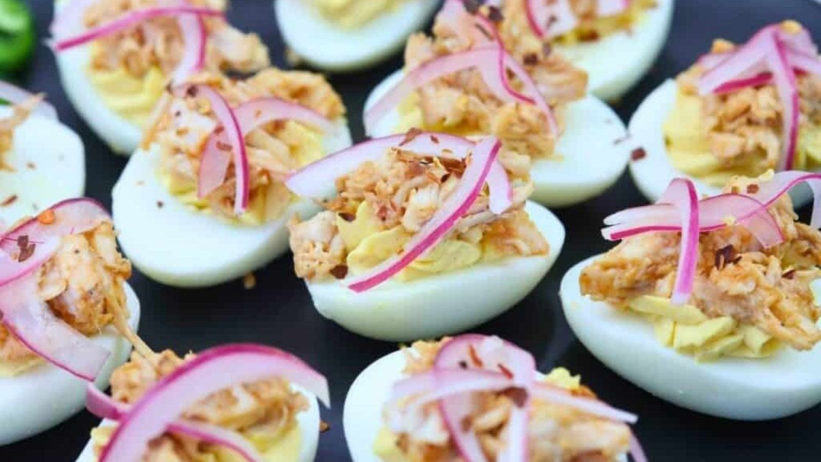 Deviled eggs topped with shredded meat and thinly sliced red onions are arranged on a black plate.