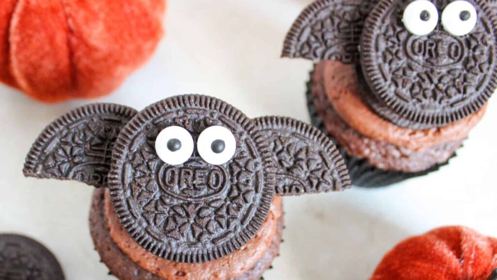 Chocolate cupcakes decorated with Oreo cookies resembling bats, complete with candy eyes. Orange pumpkins are visible in the background.