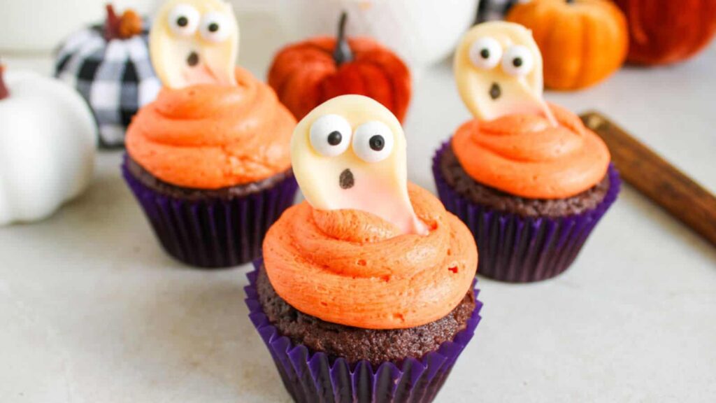 Three chocolate cupcakes with orange frosting and ghost-shaped decorations on top, arranged on a white surface with small decorative pumpkins in the background.
