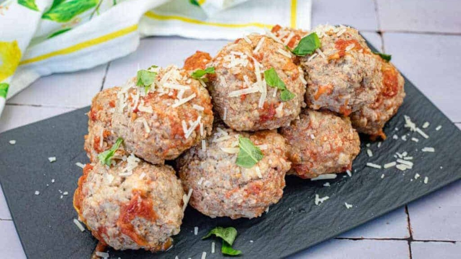 A stack of giant meatballs garnished with grated cheese and fresh herbs served on a black rectangular slate plate.