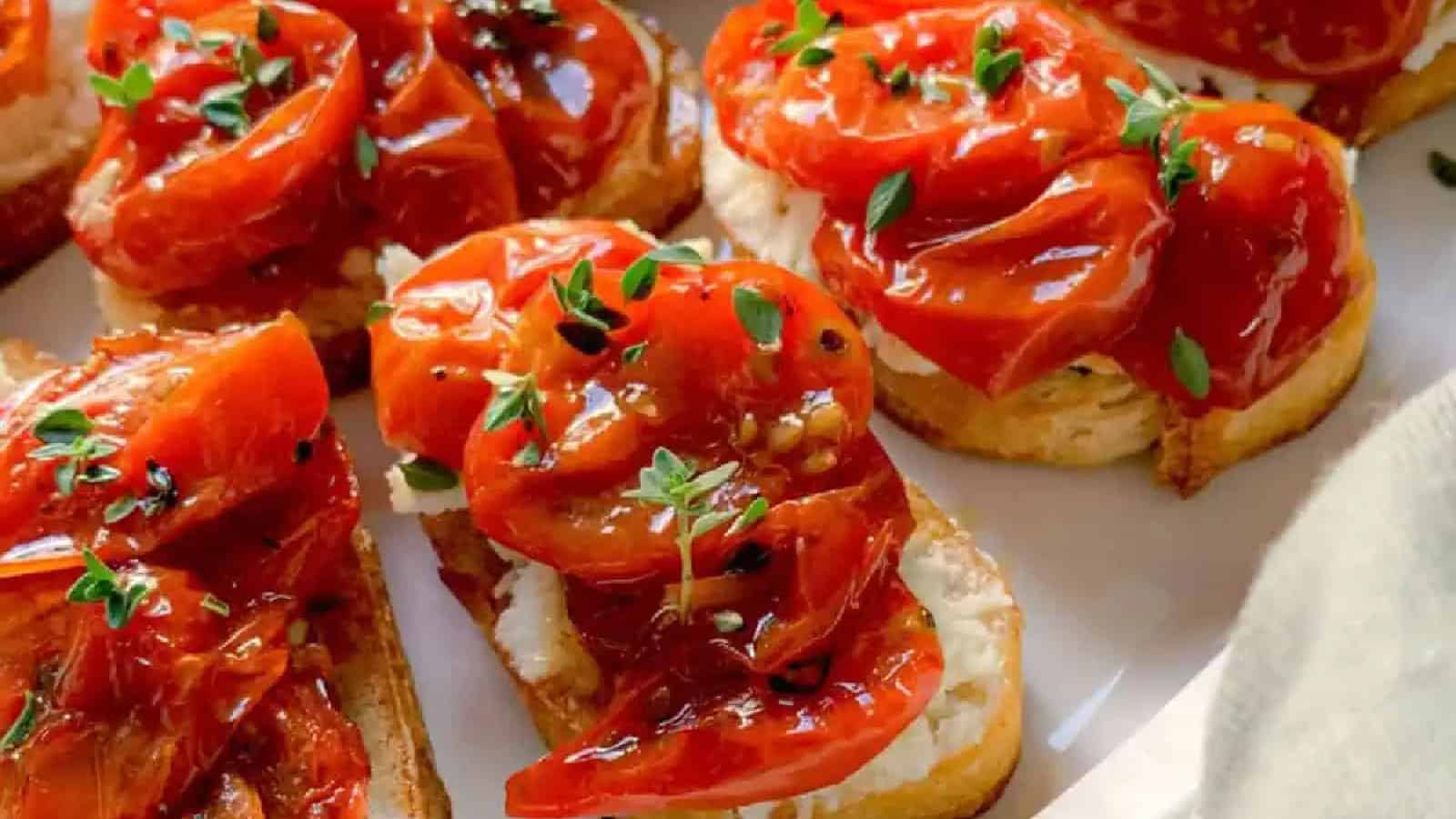 Slices of toasted bread topped with roasted tomatoes, creamy cheese, and fresh thyme on a white plate. The tomatoes are glistening, and the thyme adds a touch of green.