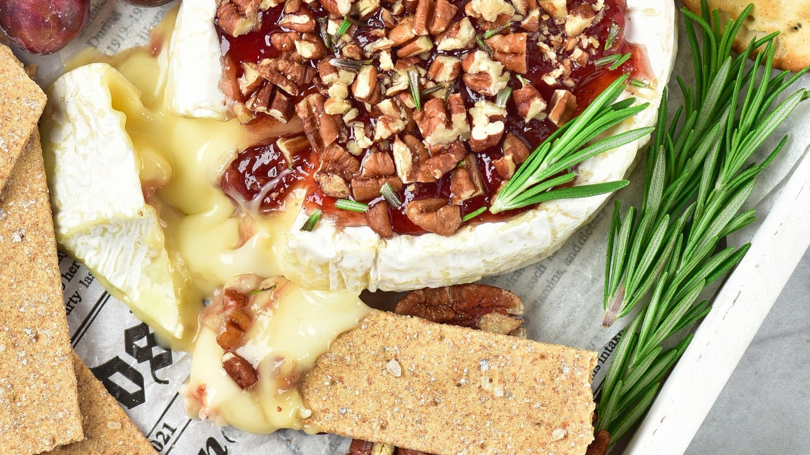 A wheel of baked brie topped with red jam, pecans, and rosemary, served with crackers and fresh rosemary sprigs.