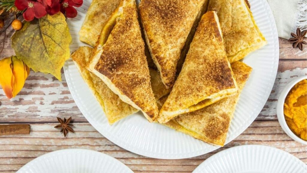 A plate of Pumpkin Turnovers arranged in a stack, with autumn-themed decorations and a small bowl of orange spread nearby.