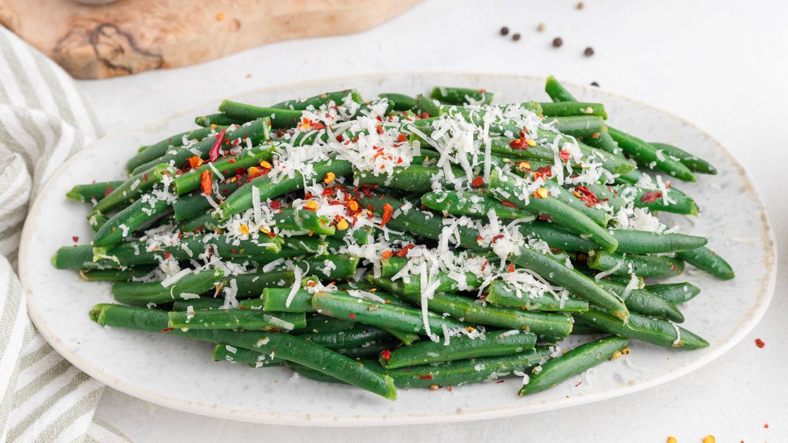 A platter of cooked green beans topped with shredded cheese, red pepper flakes, and seasonings, served on a white oval plate.