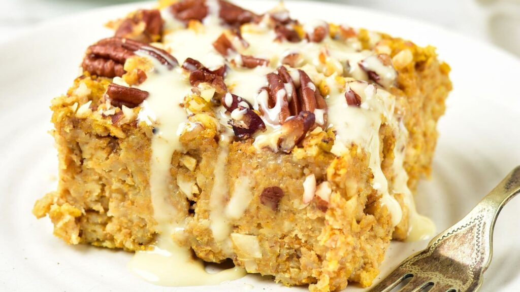 A square slice of baked pumpkin oatmeal topped with pecans and a cream-colored glaze on a plate with a fork.