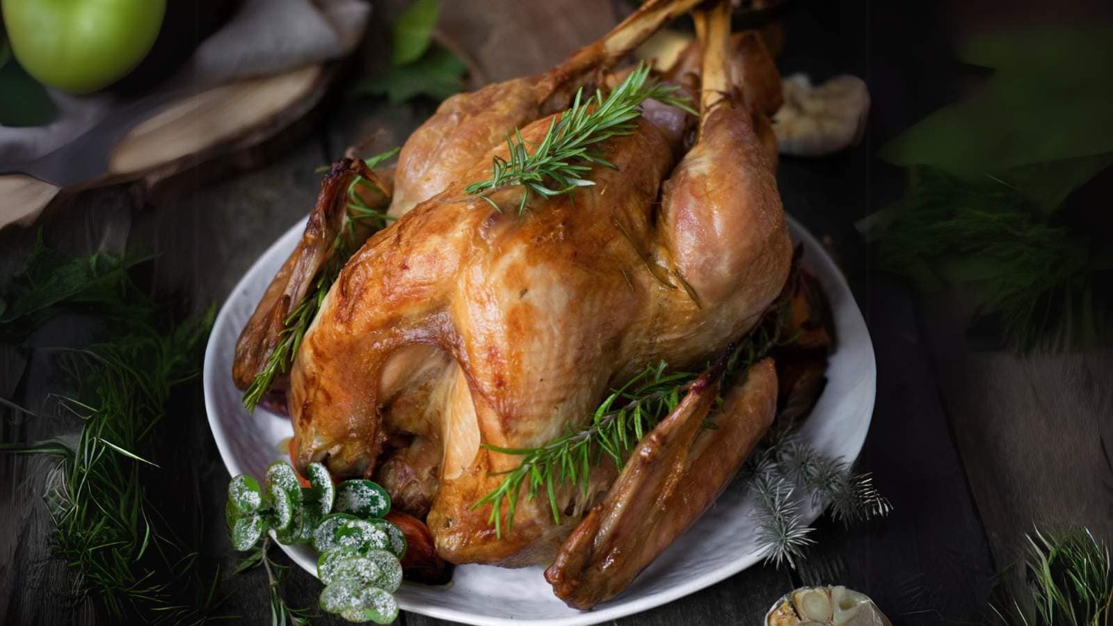 Roasted turkey on a white plate garnished with a sprig of rosemary, surrounded by herbs and sliced garlic on a rustic wooden table.