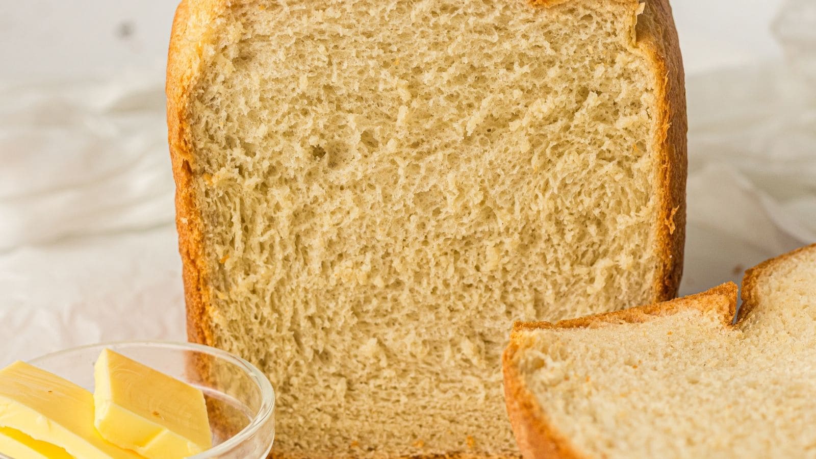 A loaf of sliced bread sits next to a glass bowl containing several pieces of butter, all placed on a white surface.